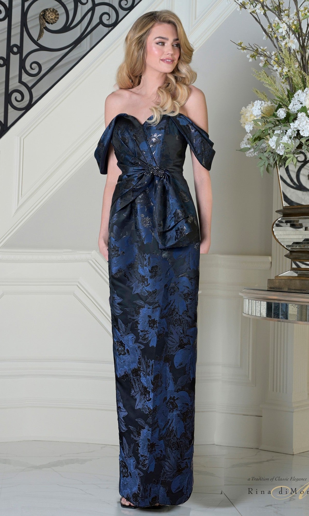 A woman stands indoors in the Long Formal Dress: Rina Di Montella RD3503, a navy off-the-shoulder, floor-length gown with floral texture and a fitted silhouette. Her hair is styled in loose waves.